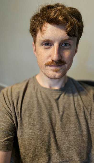 A selfie of a young man with curly reddish-brown hair, a mustache, and blue eyes, wearing a gray t-shirt.