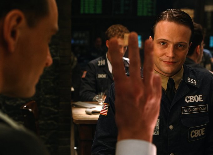 A man in a CBOE uniform labeled “G. BLOMHOLT” and number “658” smiles while holding up three fingers across a table. The caption “guy who calls volume ‘vol’” jokingly frames him as the CBOE trader who abbreviates trading volume.