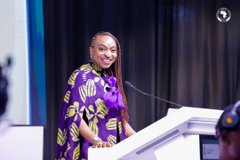 A woman wearing a purple patterned shirt standing at a white podium, likely giving a speech, with a '7' network logo visible in the top right corner.