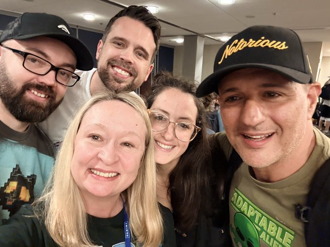 Selfie of five smiling people at an indoor event, including a man on the right wearing a black 'Notorious' cap and a green shirt reading 'ADAPTABLE ALIEN', and a man on the left wearing a black Tesla cap.