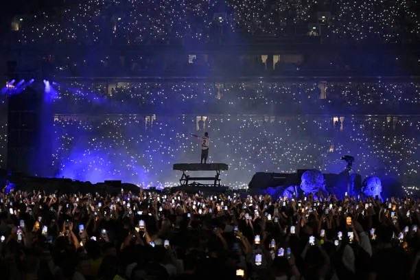 A dramatic concert photo showing a lone performer on stage in front of a massive crowd holding up illuminated phones, with a sea of light points creating a starry atmosphere under purple lighting.