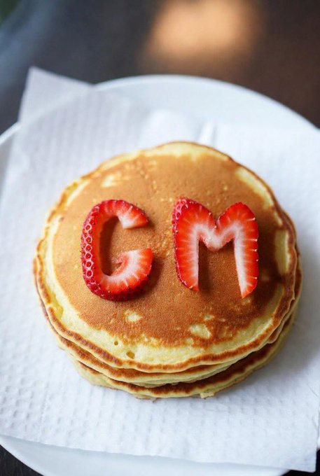 A stack of golden pancakes on a white paper napkin with strawberry slices arranged to spell out the letters 'CM' on top.