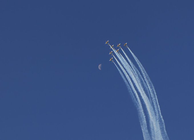 a formation of jets flying in the sky with contrails, leaving a crescent moon behind.