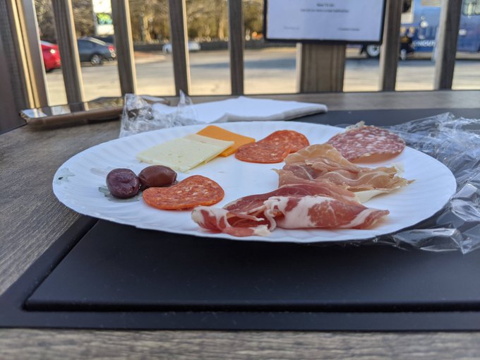 The image shows a plate of food, including cheese, meat, and olives, sitting on top of a table. The plate has been partially eaten, with only half of its contents remaining. In addition to the food items, there are also two pieces of paper visible in the scene. One is placed near the edge of the image, while the other is positioned closer to the center. The background features a view of a street and cars passing by, providing context for the location where the meal was enjoyed.