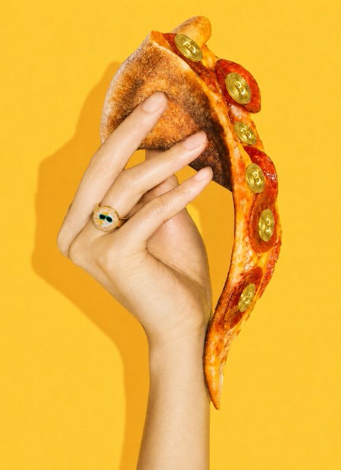 A hand wearing a gold evil-eye ring holds a slice of pizza with pepperoni and jalapeños against a vibrant yellow background in a stylized product shot.