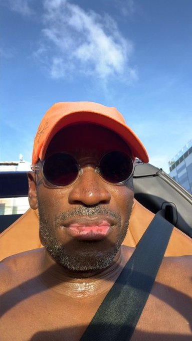 Close-up selfie of a man wearing an orange cap and round dark sunglasses, sitting shirtless in a car with the sky visible in the background.