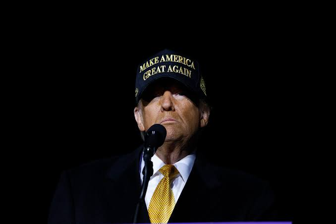 Donald Trump, wearing his signature "MAKE AMERICA GREAT AGAIN" hat, speaks at a podium against a dark background, as he prepares to meet with crypto industry leaders at the White House in what could signal significant developments for the U.S. cryptocurrency sector.