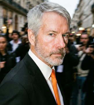 A portrait of a serious-looking, gray-haired man with a beard wearing a black suit and orange tie, with photographers and cameras visible in the blurred background.