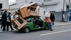 A green compact car is being loaded with furniture and a refrigerator by several workers on a city street. The scene humorously illustrates "onboarding (real)" by depicting the literal act of loading belongings into a car during a move.