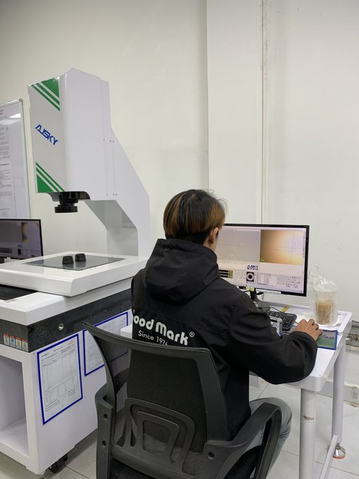 A person wearing a black 'Wood Mark Since 1976' hooded jacket sits at a desk operating an 'ALSKY' brand optical measuring or inspection machine with a monitor displaying technical data.