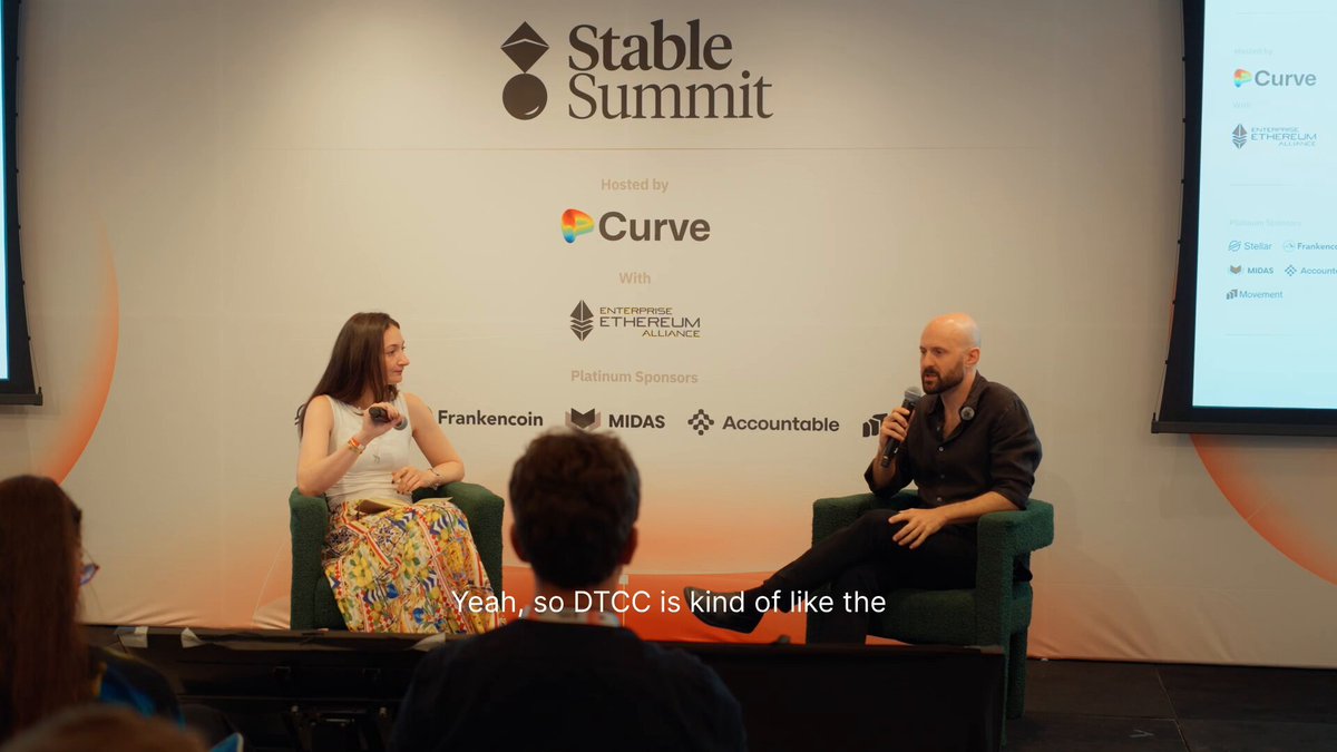The image shows a man and woman sitting at a table, with a microphone between them as they are speaking into it. The background features a white wall with various logos and text, including "@stable_summit" and "Why @The_DTCC is tokenizing on Stellar, according to @tomerweller during @stable_summit: Institutional track record Uptime and the rails to handle TradFi Trust built on years of collaboration". The image also contains a caption that reads "Focus on what is in the image and how it relates to the post."