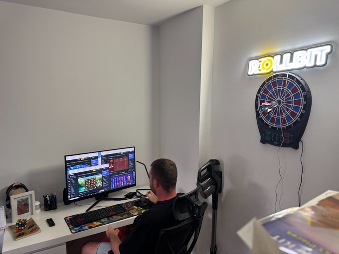 A man sitting at a desk with trading monitors in a home office featuring an illuminated 'ROLLBIT' sign and an electronic dartboard on the wall.