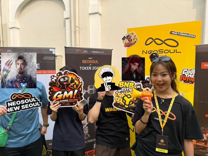 A group of four people poses at a 'NEOSOUL' booth, holding character standees and props while wearing branded t-shirts and lanyards.