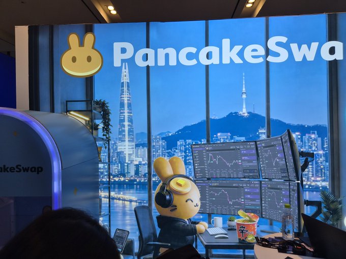 A promotional display for 'PancakeSwap' featuring their bunny mascot at a desk with multiple monitors showing trading charts, with a city skyline (including N Seoul Tower and Lotte Tower) visible through the window.