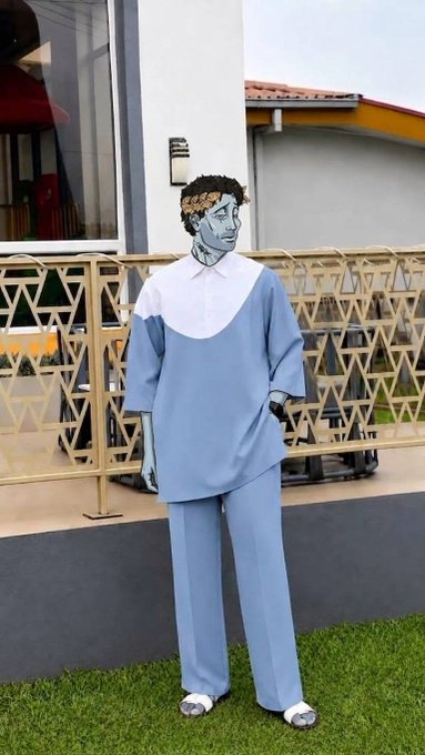 A life-sized cardboard cutout of a classical statue head wearing a laurel wreath, a light blue oversized collared shirt, and matching light blue pants stands on a grass lawn outside a building.