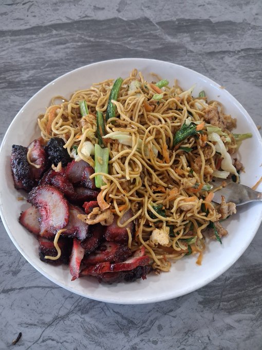 A white bowl containing Chinese-style fried noodles mixed with vegetables, served alongside sliced char siu pork on a gray stone-patterned countertop.