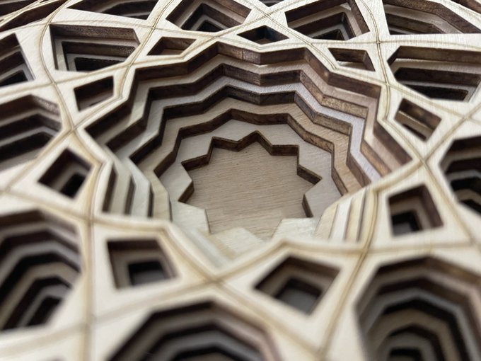 A geometric wooden structure with intricate laser-cut patterns creating an optical illusion of depth, featuring a star-shaped opening in the center.