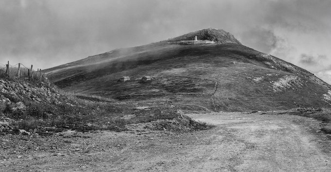 A black-and-white landscape photograph of a misty mountain with a winding dirt path leading toward a small structure at the summit.