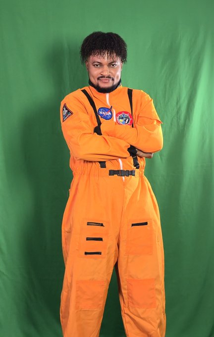 A man with curly hair posing with arms crossed in an orange NASA astronaut jumpsuit against a green screen.