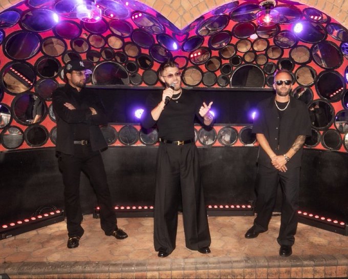 Three men dressed in all-black outfits posing on stage with a microphone, with one wearing a NY Yankees cap and sunglasses, against a backdrop of circular red-lit decorative elements.
