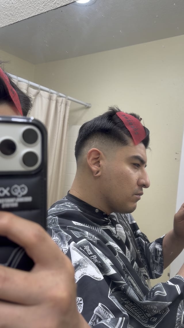 A man in a black barber cape takes a mirror selfie while styling his fresh fade haircut, with red comb guides still in place on top of his head, capturing the work-in-progress that mirrors the "practice makes perfect" mantra shared in the post.