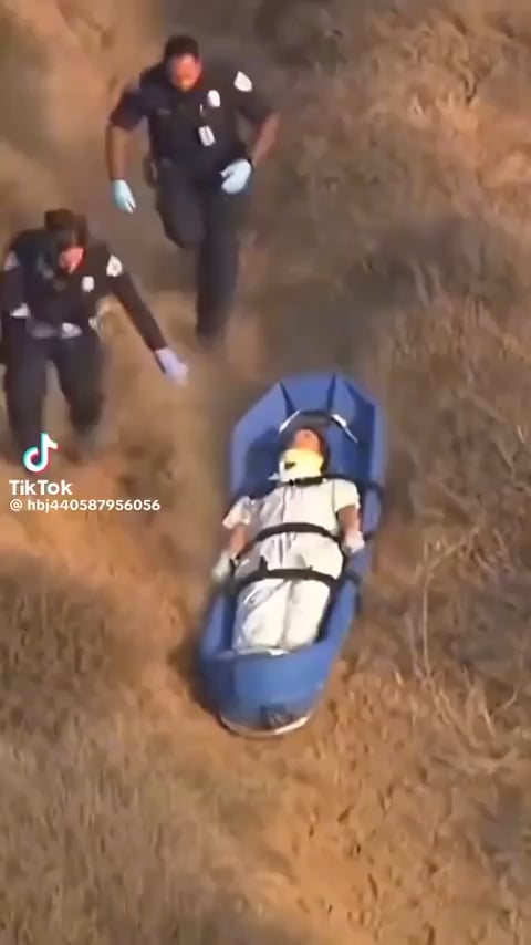 The image shows a person lying on their back, possibly injured or unconscious, being attended to by two police officers who are running towards them with medical equipment. One of the officers has a blue stretcher attached to his back. The scene takes place in an open field, and there is also text visible that reads "BTC Lately".