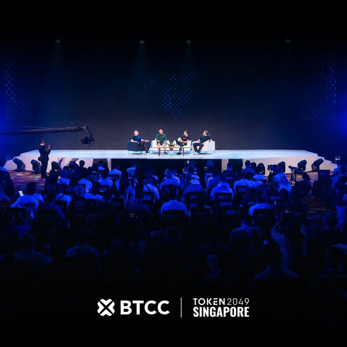 A panel discussion takes place on a brightly lit stage at TOKEN2049 Singapore, hosted by BTCC, with five seated speakers engaged in conversation while a camera crew films the event and a large audience watches from the darkened auditorium below; the overlaid text reads "BTCC | TOKEN2049 SINGAPORE," tying the image to the company's celebration of 15 years in crypto and their "0-Barrier Trading" campaign promoting 0 fees, 0 friction, and 0 panic at their Marina Bay Sands booth.