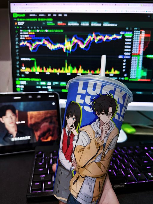 A hand holding a 'LUCK' branded cup featuring anime-style characters, with a computer monitor showing cryptocurrency trading charts and a tablet displaying a video in the background.