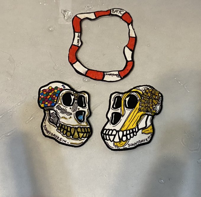 Three hand-drawn stickers or patches, two skull designs with the text 'sunnydayz.eth' and a red and white striped circular design with 'BAYC', rest on a wet surface.