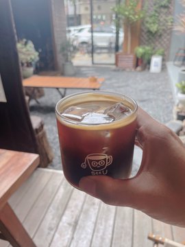A hand holds a clear glass of iced coffee with the text 'crul' on it, set on a wooden deck overlooking a courtyard with wooden benches and potted plants.