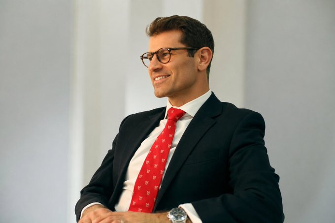 The image features a man wearing glasses, a suit, and a red tie. He has his hands folded together in front of him as he looks off to the side with a smile on his face. The background appears to be a white wall or a gray surface, which contrasts nicely with the man's attire. The caption below the image reads "Giga Gilbert with huge $QNT news. Well done @quantnetwork".