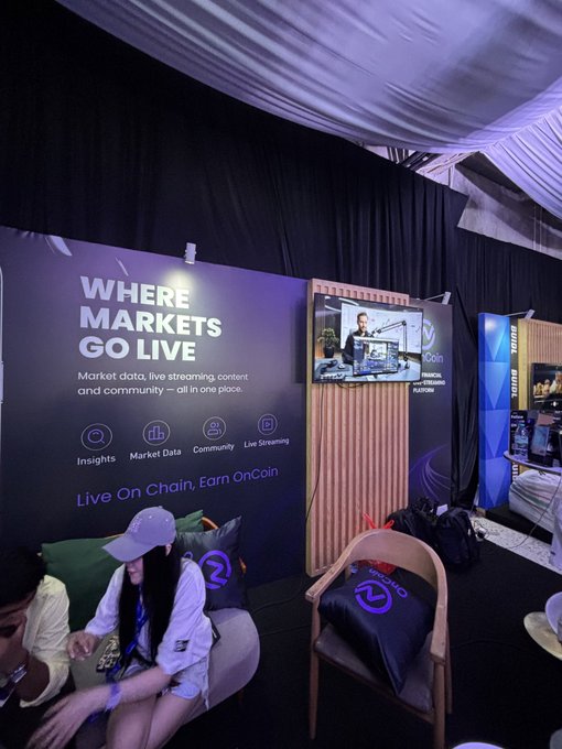 A promotional booth for OnCoin Live at an event, featuring a large banner reading "WHERE MARKETS GO LIVE - Market data, live streaming content and community – all in one place. Insights, Market Data, Community, Live Streaming. Live On Chain, Earn OnCoin," alongside a TV screen showing what appears to be a live broadcast. A woman in a white cap sits in the booth area, with branded merchandise visible on nearby chairs, showcasing OnCoin Live's offerings of integrated market insights and community features.