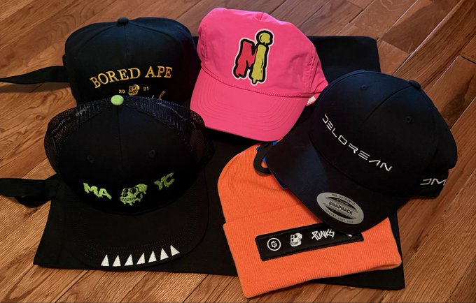 A collection of four snapback hats rests on a wooden floor, including a black hat reading 'BORED APE', a pink hat with 'N1', a black hat with 'JELOREAN', and an orange Stüssy beanie.