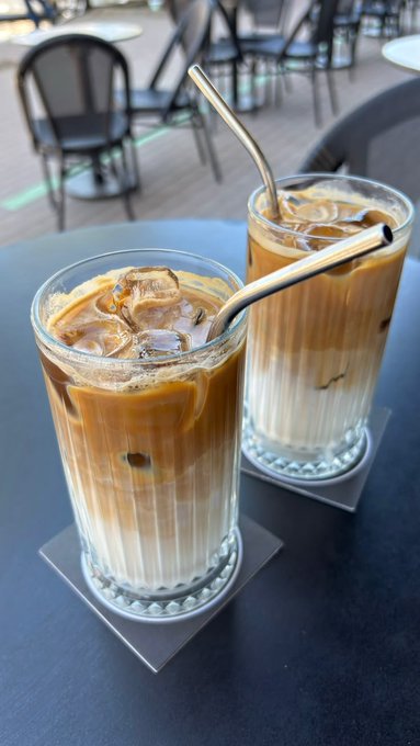 Two tall glasses of iced coffee with metal straws resting on coasters on a dark outdoor cafe table.