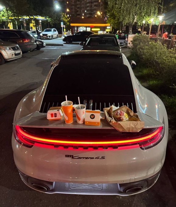 A white Porsche 911 Carrera 4S parked at night with its rear spoiler raised, displaying McDonald's drinks, food boxes, and a wrapped burger on the back.