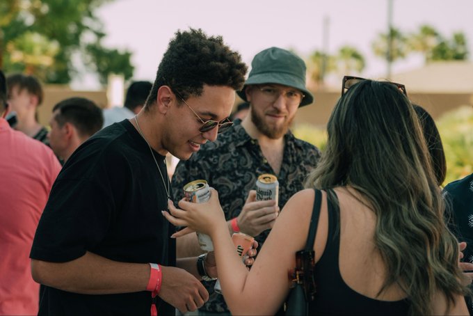 Two young men at an outdoor social event holding cans of beer, one wearing a bucket hat and floral shirt, the other with curly hair and round sunglasses, while a woman with a bottle of beer stands in the foreground.