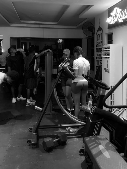 A black and white photo inside a gym showing several people, with a woman in the foreground working out on a machine; a neon sign partially reads 'Beau' and text on the wall reads 'DON'T STOP WHEN YOU'RE TIRED'.