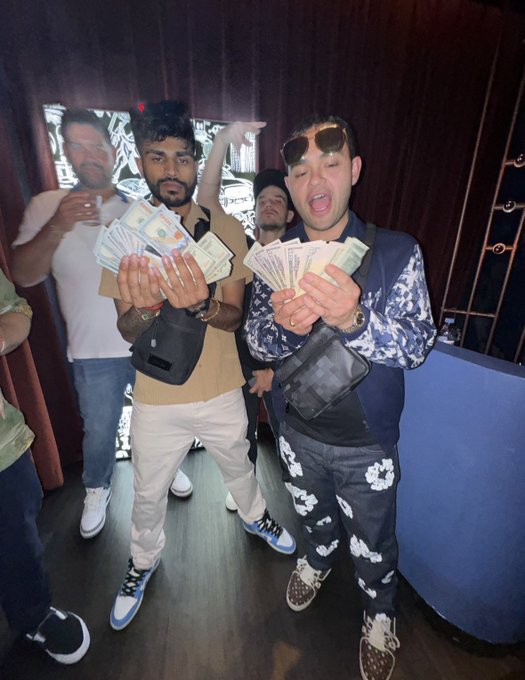 A group of men in a club-like setting pose for a photo, with two in the foreground holding up fans of US dollar bills and wearing designer outfits including a Gucci-patterned jacket.