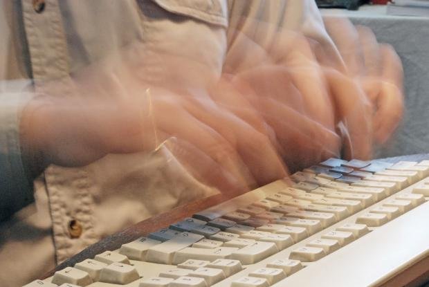 A motion-blurred photo of a person's hands typing rapidly on a white computer keyboard, wearing a beige button-up shirt.