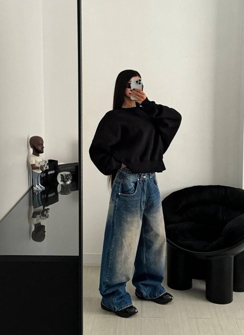 A mirror selfie of a woman wearing an oversized black sweatshirt and baggy blue jeans, standing in a room with a black chair and a small sculpture.