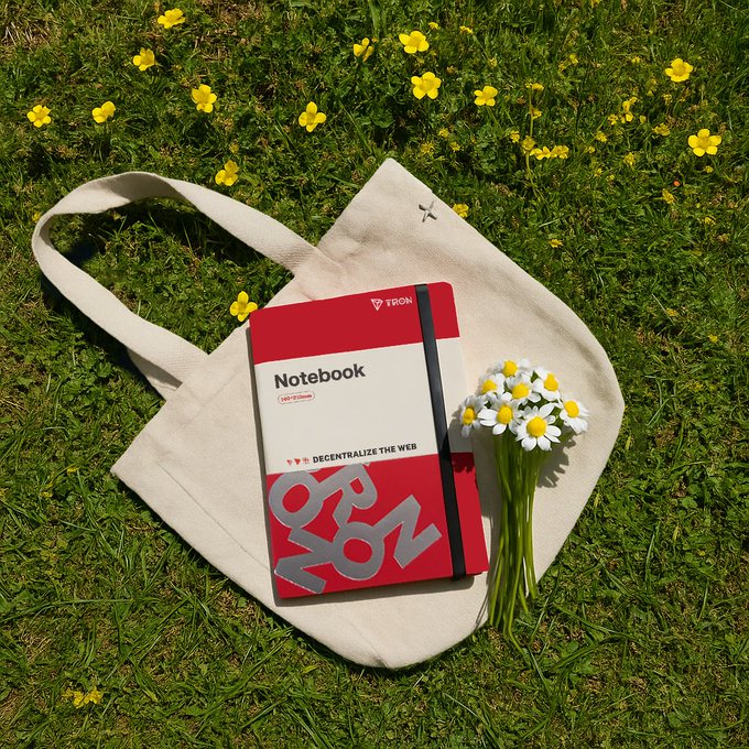 A red TRON 'Notebook - Decentralize the Web' with 140*210mm dimensions resting on a beige canvas tote bag, accompanied by a small bouquet of daisies, set against a grassy background with yellow buttercup flowers.