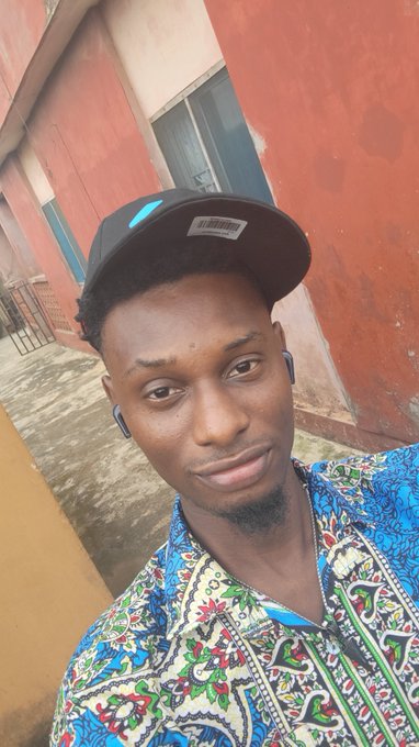 A young man wearing a black baseball cap and a colorful patterned shirt smiles for a selfie outside a building with a red wall.
