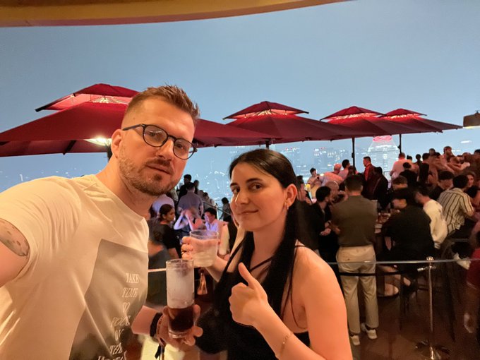 A couple poses for a selfie at a crowded, umbrellas-lined rooftop event at dusk, with the woman giving a thumbs-up while holding a drink, capturing the social atmosphere of the TOKEN2049 party where Cardano ($ADA) is the main topic of discussion.