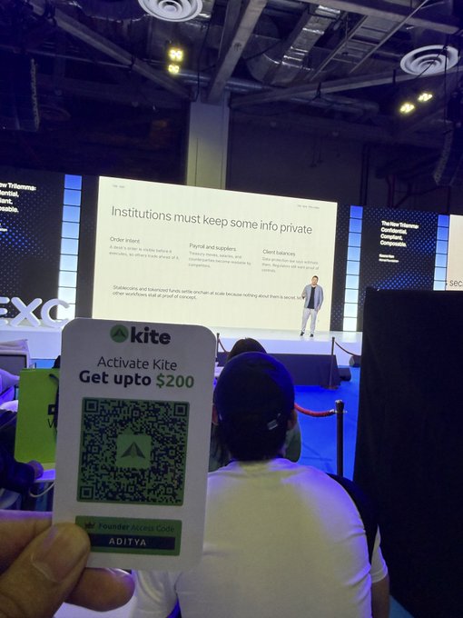 Photo taken at a conference showing a hand holding a "kite" QR code card labeled "Activate Kite, Get upto $200" with Founder Access Code ADITYA, in front of a stage displaying "Institutions must keep some info private."