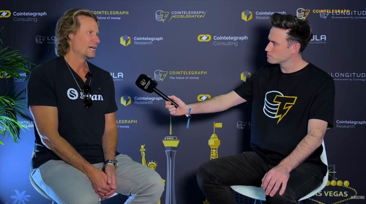 The image features two men sitting down, one of whom appears to be a PR representative for Bitcoin. They are engaged in conversation with each other, possibly discussing the future of Bitcoin or another digital asset. The background includes a black wall adorned with various logos and text, which could be related to the Bitcoin PR machine's activities.