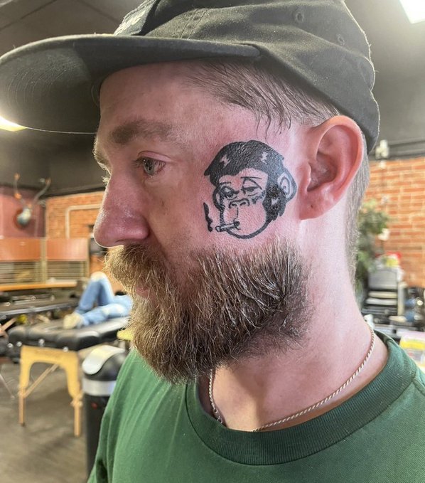 A bearded man in a green shirt and backward cap has a small black line-art portrait tattooed on his cheek. The image accompanies the post “nothing will ever compare to what nfts did to mfers,” evoking an NFT-style customized face.