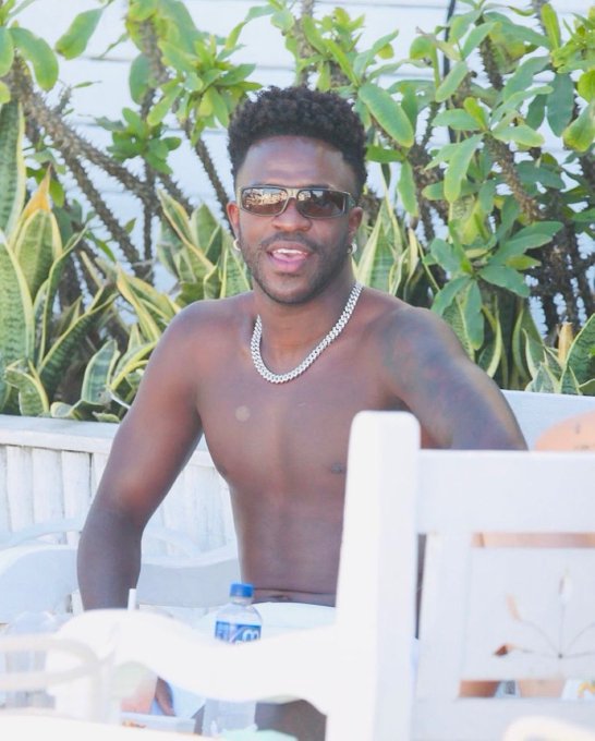 A shirtless man, likely Vini Jr., sports a new look with a fresh haircut and stylish sunglasses while relaxing outdoors, accessorized with a silver chain necklace against a backdrop of lush greenery.