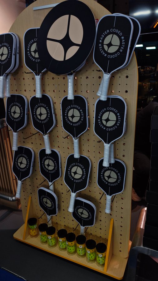 A retail display features multiple black pickleball paddles branded with 'ASTER CODE' and a star logo hanging on a pegboard, with small yellow smiley-face jars lined up on the bottom shelf.