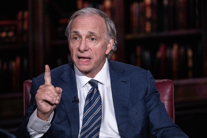 A gray-haired man in a blue suit and striped tie gestures emphatically while speaking in front of a bookshelf backdrop. The image accompanies a quote attributed to Ray Dalio about pursuing ambitious goals, suggesting this is him making the statement.