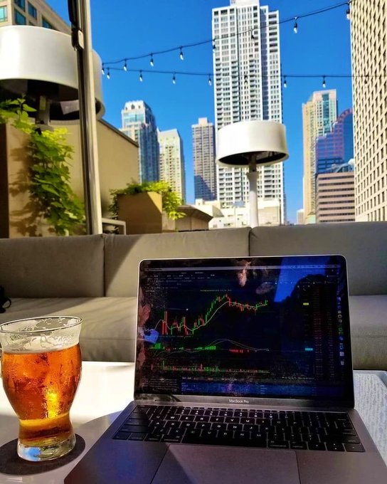 A MacBook Pro on an outdoor patio displaying trading charts with a glass of iced tea nearby, overlooking a city skyline with tall buildings under a clear blue sky.